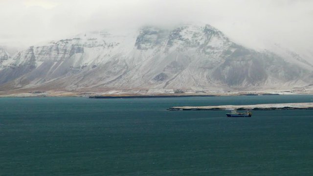 Extreme long shot view of a ship sailing in sea water with snowy Esja mountain in background