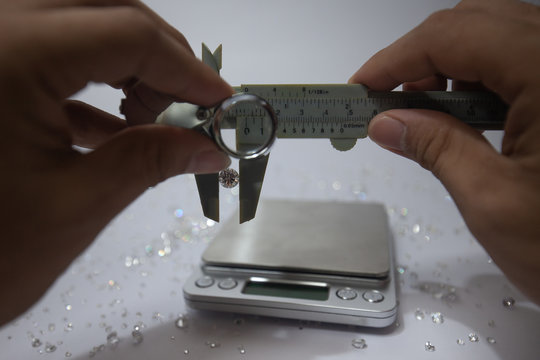 Hand holding a diamond with weight scale and vernier caliper, tweezers and a pile of diamond. Worker measures the weight of gems on a jewelry scale isolated on white background. - Powered by Adobe