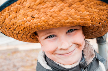 Close up portrait of a sweet smiling light-skinned boy with blue eyes and a dirt-smeared face, wearing a straw hat with a large brim. A dirty little farmer.