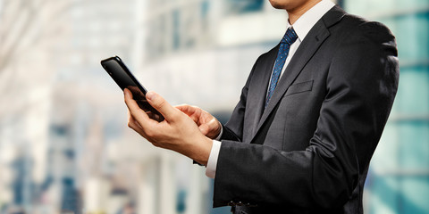街角に立ち、スマートフォンを操作するビジネスマンのイメージ素材