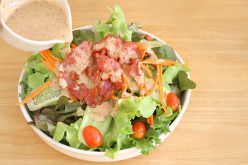 Close up fresh vegetable salad (lettuce, carrot, cucumber, tomato)  with fried bacon and pour by roasted sesame Japanese dressing in white bowl on wooden table background