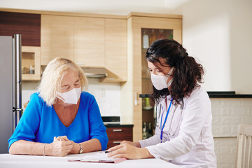 Young female doctor in medical mask showing prescription to senior patient