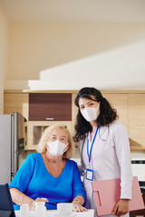 Portrait of positive senior woman in medical mask and doctor visiting her at home