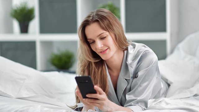 Adorable young smiling woman in pajamas use smartphone lying on bed. Medium shot on RED camera