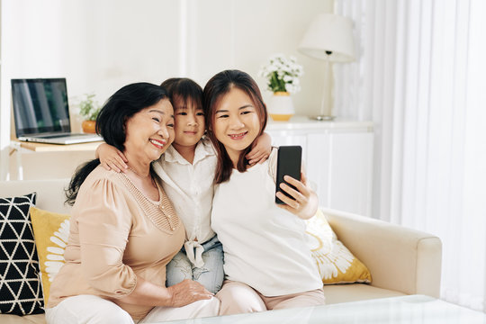 Cheerful young Vietnamese woman taking selfie with her aged mother and little daughter at home