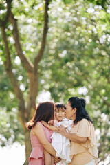 Fototapeta premium Beautiful granny and mother hugging and kissing little girl when standing in park