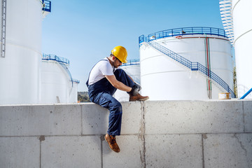 Handsome Caucasian workman in overalls and with helmet on head sitting on wall and tying shoelace. In background are oil tanks. Refinery exterior.