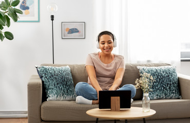 people, technology and leisure concept - happy smiling young african american woman in headphones sitting on sofa and listening to music or watching movie on tablet pc computer at home