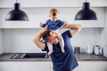 Young proud smiling positive single dad playing holding his beloved son on shoulders and having fun and kitchen. Toddler holding mixing spoon and playing with his dad.