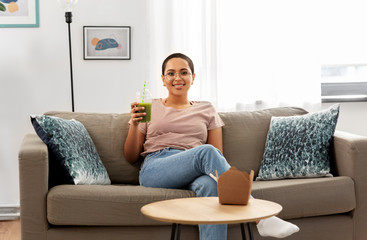 people, eating and leisure concept - happy smiling african american young woman sitting on sofa and drinking smoothie from plastic cup with paper straw at home
