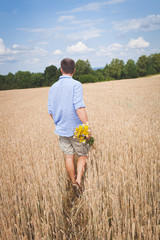 Naklejka na ściany i meble Счастливая мужчина с букетом диких цветов прогуливается в пшеничном поле. Молодой человек с цветами и пшеничное поле на фоне голубого неба. Вид с зади на мужчину. Концепция счастья и образ жизни