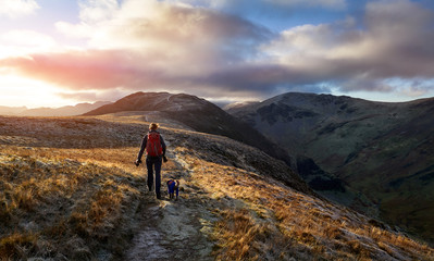 A hiker and their dog walking towards the mountain summit of High Spy from Maiden Moor at sunrise on the Derwent Fells in the Lake District, UK.