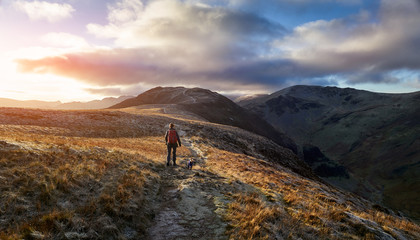 A hiker and their dog walking towards the mountain summit of High Spy from Maiden Moor at sunrise on the Derwent Fells in the Lake District, UK.