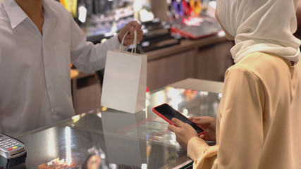 An upwardly mobile Asian Muslim woman using a mobile phone - smartwatch to pay for a product at a sale terminal with nfc identification payment for verification and authentication