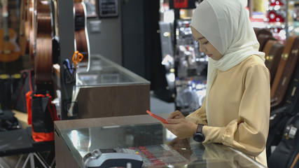 An upwardly mobile Asian Muslim woman using a mobile phone - smartwatch to pay for a product at a sale terminal with nfc identification payment for verification and authentication