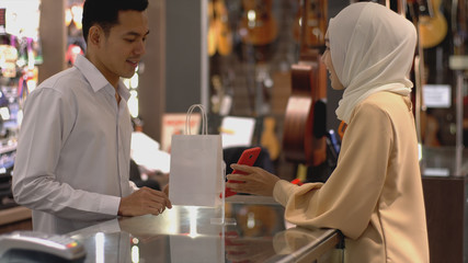 An upwardly mobile Asian Muslim woman using a mobile phone - smartwatch to pay for a product at a sale terminal with nfc identification payment for verification and authentication