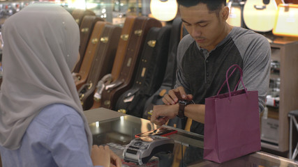 An upwardly mobile Asian Muslim man using a mobile phone - smartwatch to pay for a product at a sale terminal with nfc identification payment for verification and authentication