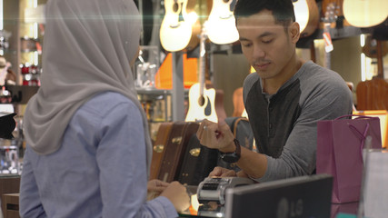 An upwardly mobile Asian Muslim man using a mobile phone - smartwatch to pay for a product at a sale terminal with nfc identification payment for verification and authentication
