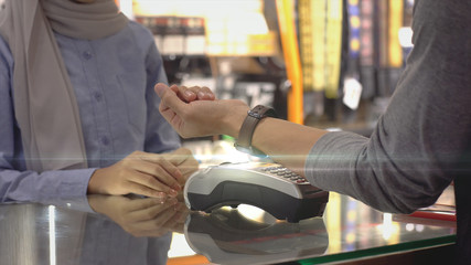 An upwardly mobile Asian Muslim man using a mobile phone - smartwatch to pay for a product at a sale terminal with nfc identification payment for verification and authentication