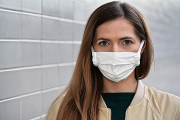 Portrait of young woman with home made cotton face mouth virus mask, grey tiles wall in background,...