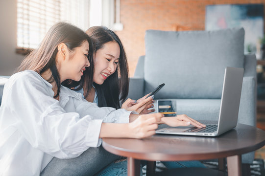 Asian Women Friends Searching For Online Shopping In Smartphone And Laptop While Holding Credit Card In Living Room