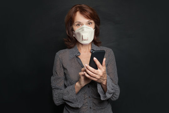 Mature business woman in medical mask using smartphone on black background
