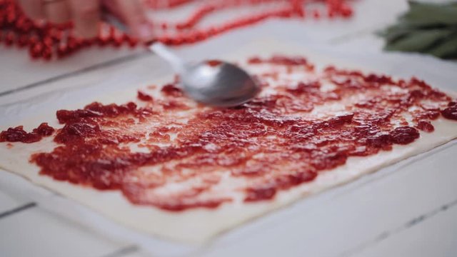 Spreading red jam on a puff pastry dough sheet with a spoon. Handheld shot.