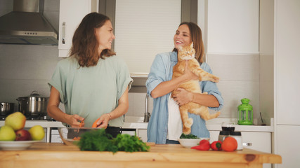 Happy lesbian couple preparing dinner in the kitchen, time together, one wife holds a red ginger cat in her arms