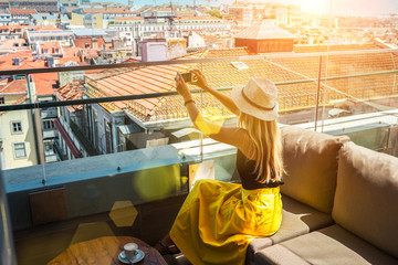 Young traveler woman with smartphone sitting in rooftop cafe Lisbon and takes pictures of city...
