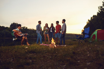 A group of people sitting by the bonfire next to the tent at night in the summer in autumn.