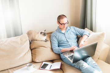Young man is siiting on the floor and working at home on a computer with the phone, papers, cat and a cup of coffee. Remote work, work from home.