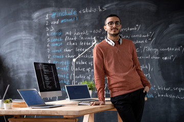 Portrait of bearded Arabian software developer with wireless headphones around neck standing at desk with computers in office