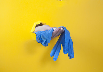 Female hand holds blue rubber gloves on a yellow background. Hand in the hole. Hygiene. Purity. covtd-19