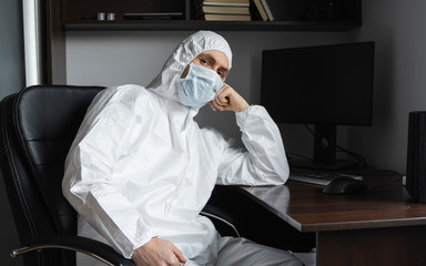 Man in medical mask and protective suit, rubber gloves sits at home and works with pc at the table during quarantine. Designer, artist, architect, businessman at remote work in a pandemic covid.