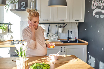 Happy cute blonde phoning and making dinner in bright kitchen