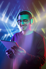 Excited young Arabian man addicted to gadget listening to music in headphones playing online game against bright geometric background