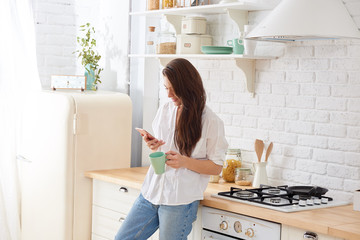Young beautiful woman using cell phone and having a coffee in the kitchen