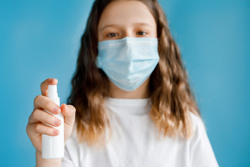 A young girl in a medical protective mask holds an antiseptic bottle in her hands and looks into the frame