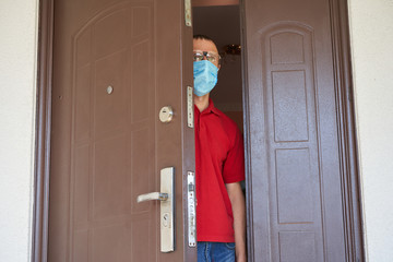 masked man looks out the door,a man in a red shirt and medical mask looks out through the slit of...