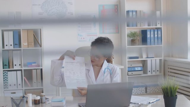 Medium shot of female doctor wearing medical gown sitting at desktop at computer and showing interlocutor how to fill in form via internet connection