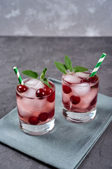 Fresh cherry lemonade with ice, mint and paper straw in sparkling glasses on gray table background, copy space. Cold summer drink. Berry cocktail