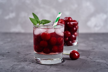 Fresh cherry lemonade with ice, mint and paper straw in sparkling glasses on gray table background, copy space. Cold summer drink. Berry cocktail