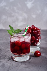 Fresh cherry lemonade with ice, mint and paper straw in sparkling glasses on gray table background, copy space. Cold summer drink. Berry cocktail
