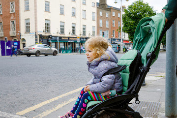 Cute little toddler girl sitting in baby stroller while walking with dad on the streets of big...