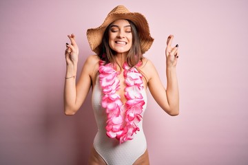 Young beautiful brunette woman on vacation wearing swimsuit and Hawaiian flowers lei gesturing finger crossed smiling with hope and eyes closed. Luck and superstitious concept.