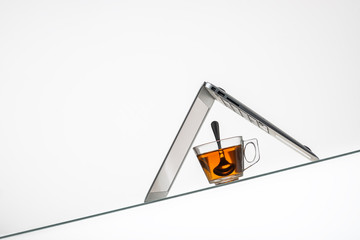 cup with hot herbal tea and laptop on inclined glass table surface 