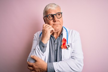 Fototapeta premium Senior handsome hoary doctor man wearing stethoscope and red HIV ribbon on coat with hand on chin thinking about question, pensive expression. Smiling with thoughtful face. Doubt concept.
