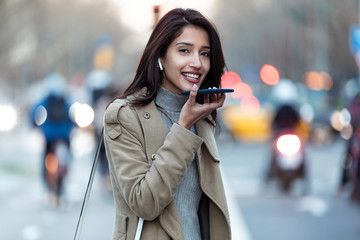Pretty young woman using voice recognition system on her smartphone while standing in the street.