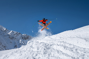 Naklejka na ściany i meble Male athlete skier in an orange trigger makes a jump trick with flying snow powder against the backdrop of snow-capped Caucasus mountains and a blue sky. Winter Extreme Sports Concept