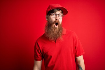 Young handsome delivery man wearing glasses and red cap over isolated background In shock face,...